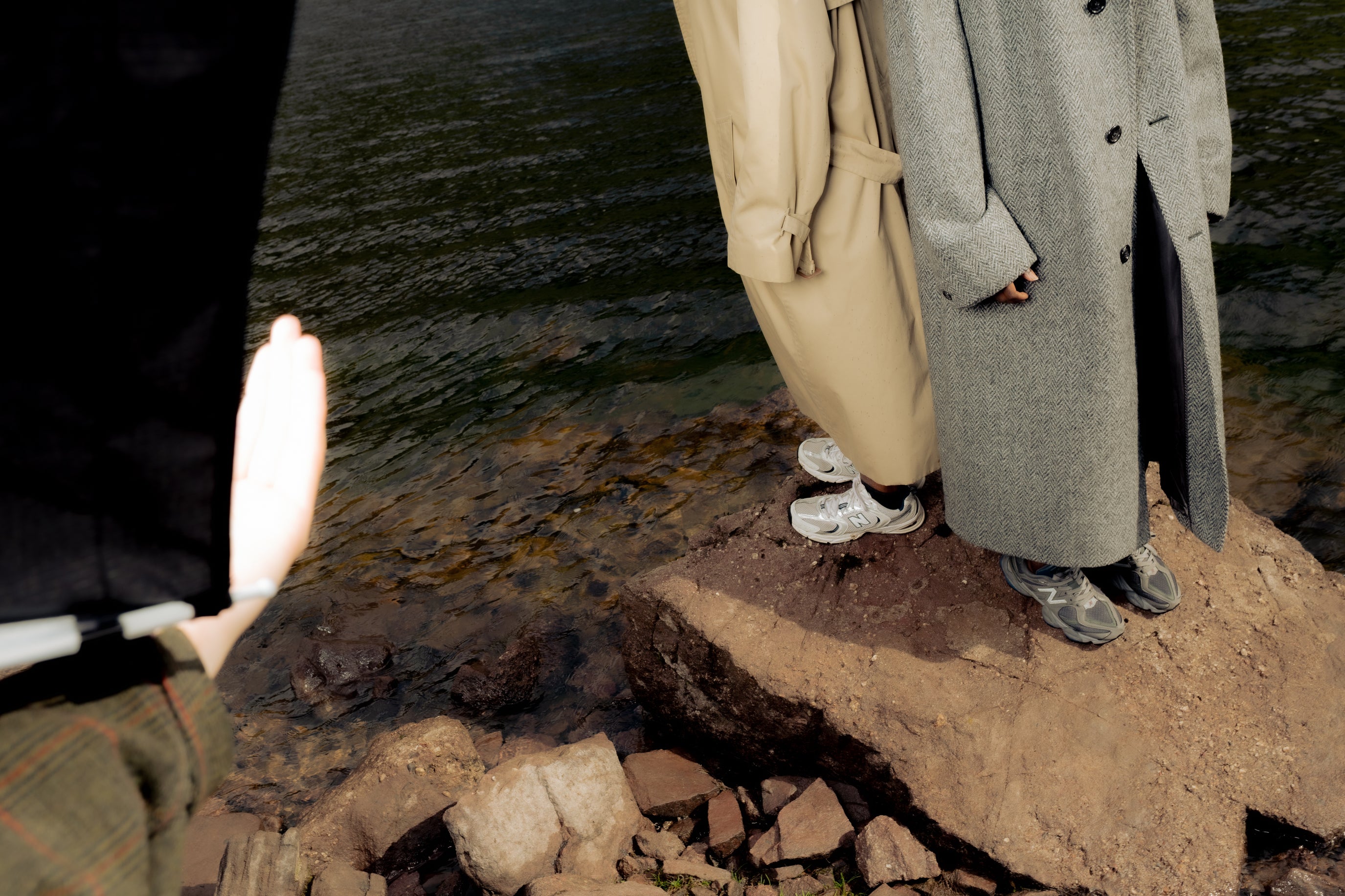 Editorial detail of two figures in long coats and sneakers standing on rocks beside Welsh water Maison Nwar fashion photography.