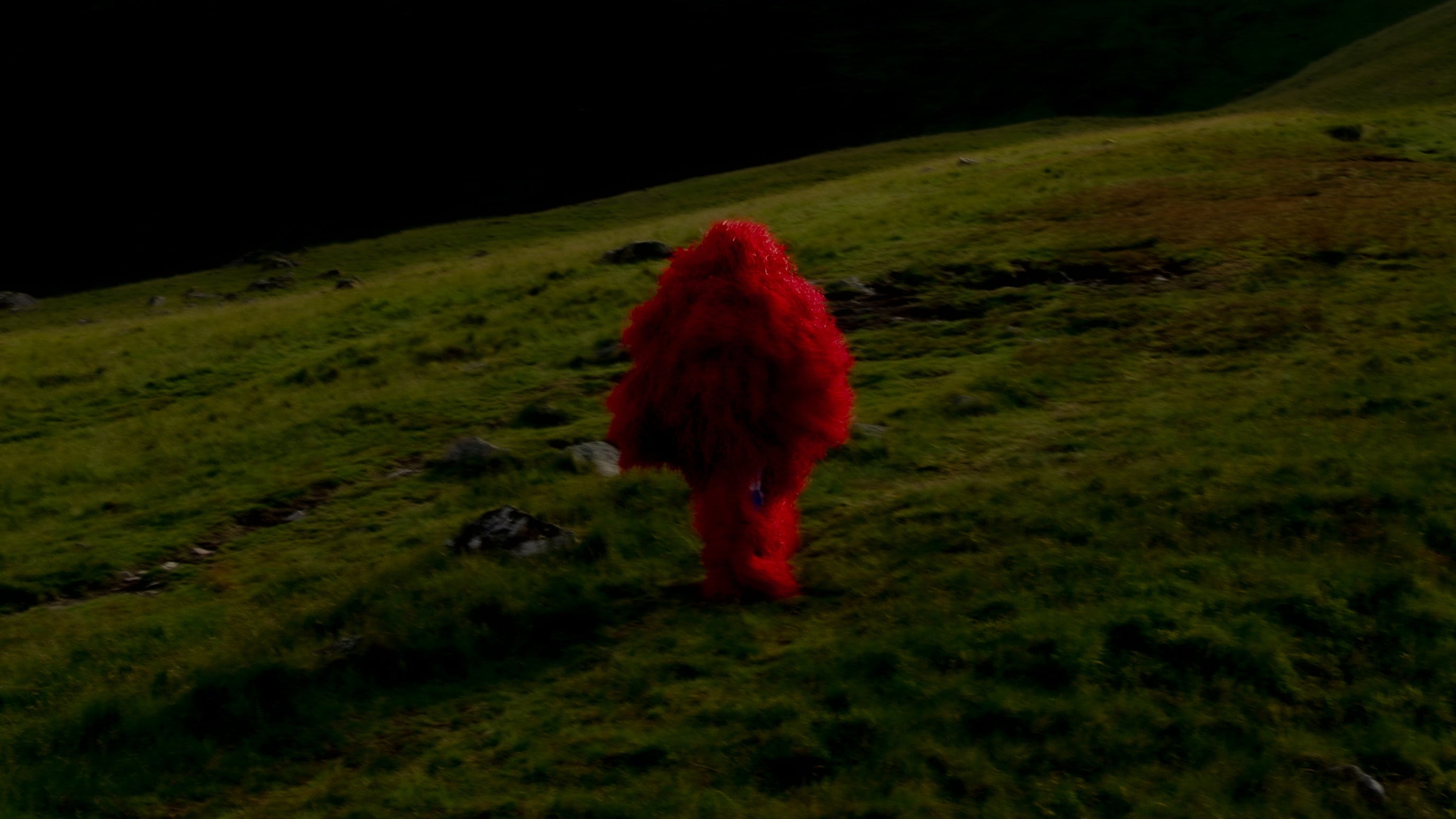 The Kankurang figure in a red costume walking alone across the grassy Welsh hills under a dark sky Maison Nwar cultural editorial.