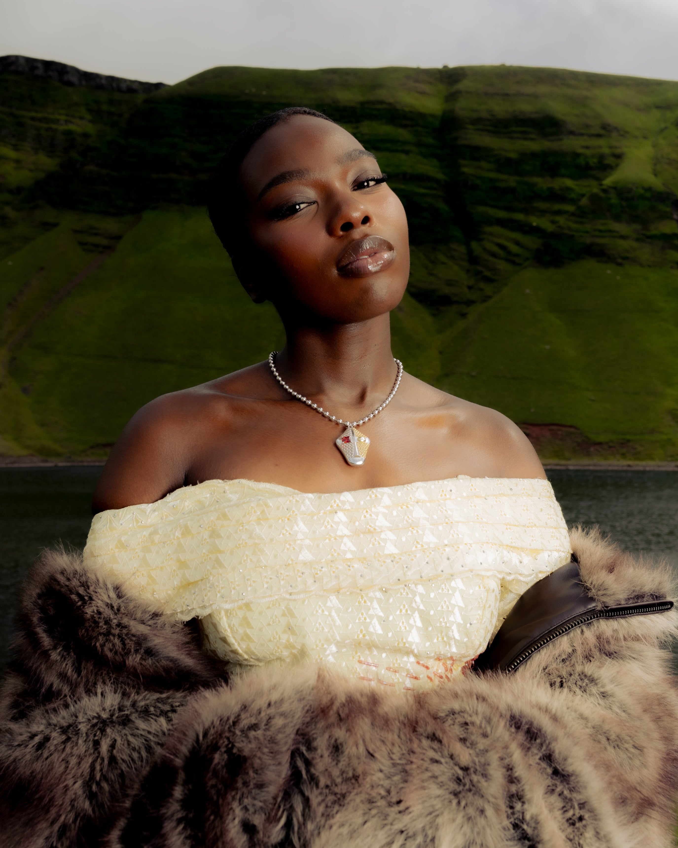 Senegalese woman in a white dress and fur wrap wearing a Maison Nwar sculptural silver pendant necklace with red stone, photographed in the Welsh mountains.