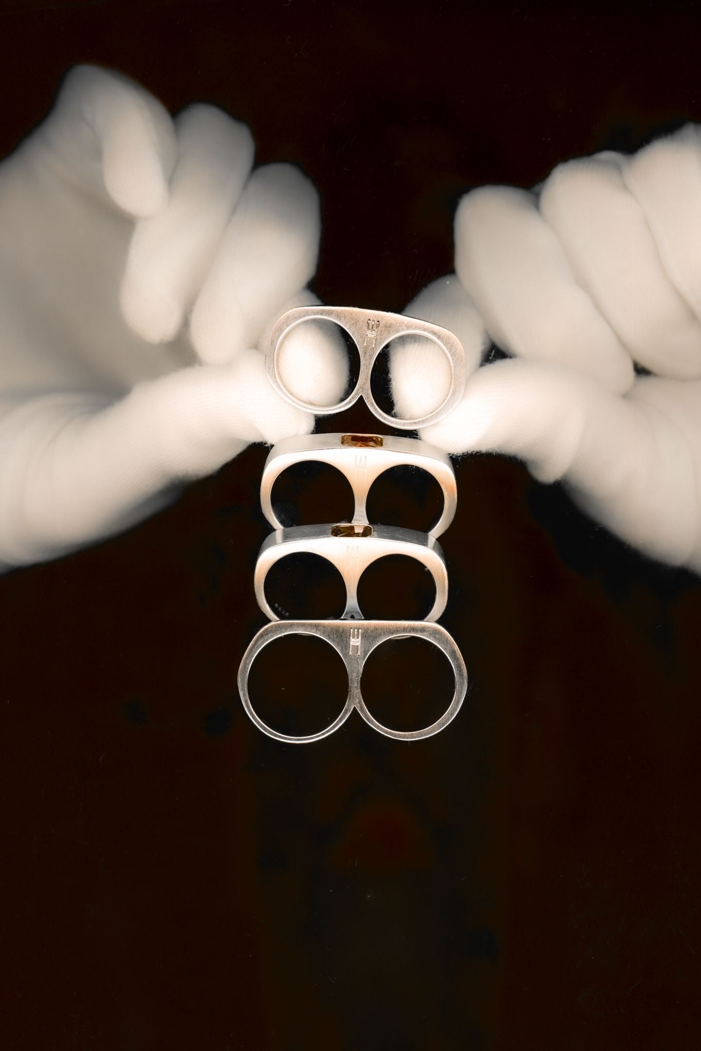 White-gloved hands holding a vertically stacked set of silver double-ring pieces against a dark red background, arranged symmetrically to emphasize form and repetition.