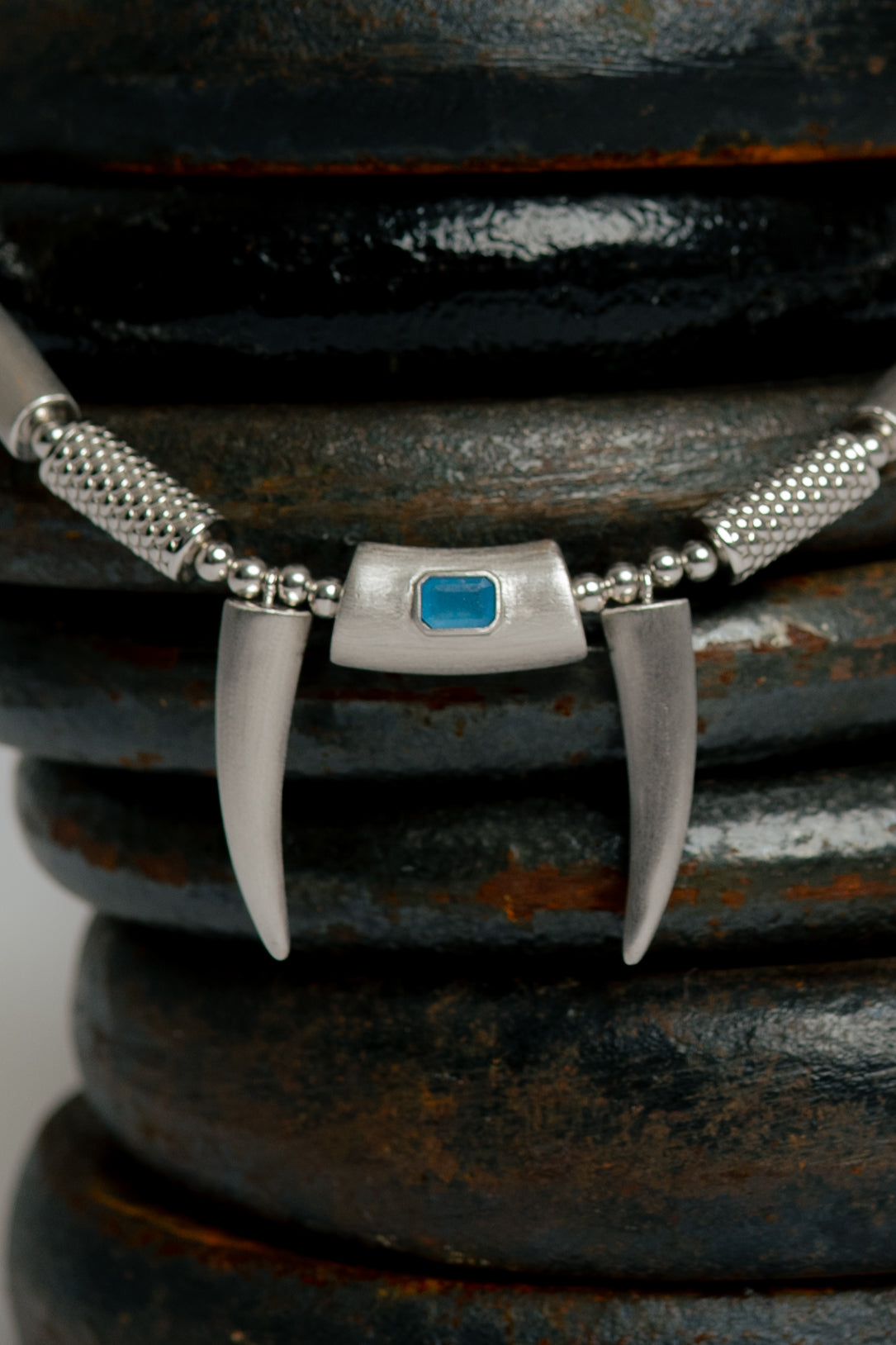 Close-up of a silver necklace with a blue stone and claw-like pendants on a dark textured surface.
