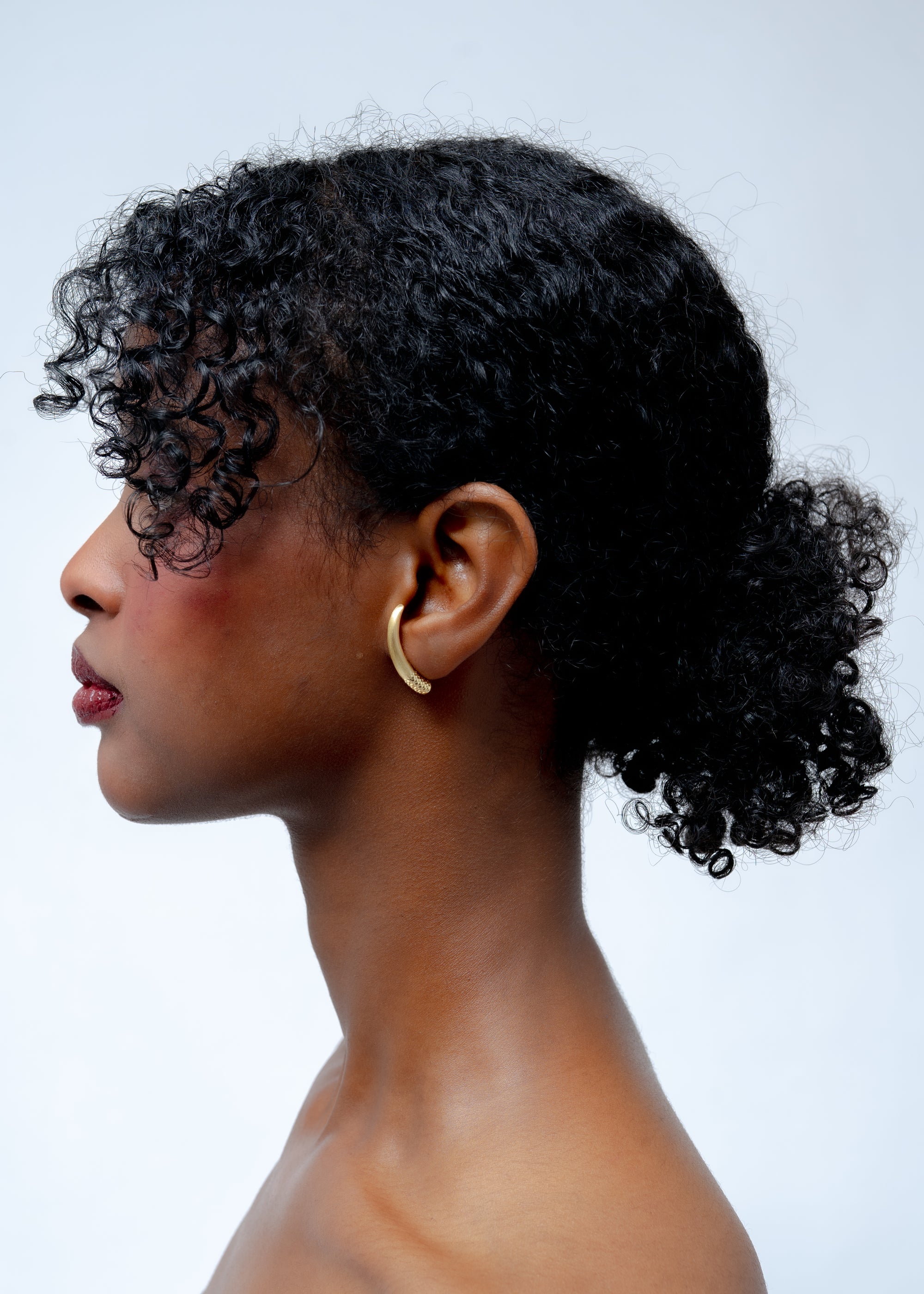 Side profile of a person wearing a small gold hoop earring against a light background.