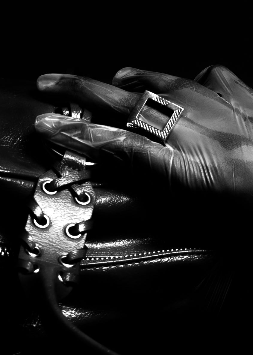 Close-up of a gloved hand wearing a square silver ring on a black leather surface.