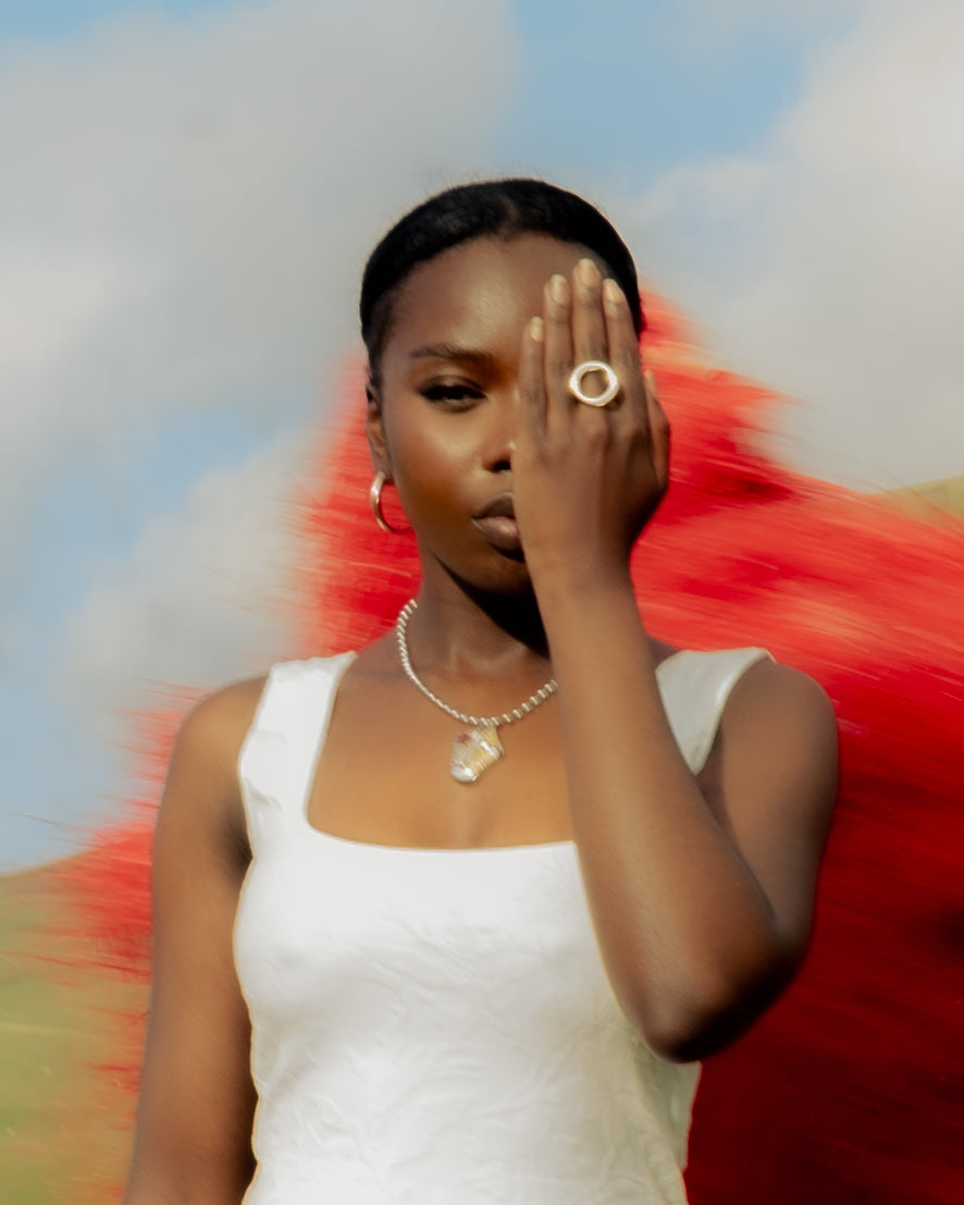 Close-up of a Senegalese woman in a white dress wearing Maison Nwar jewelry, covering one eye with her hand red Kankurang figure blurred in the background.