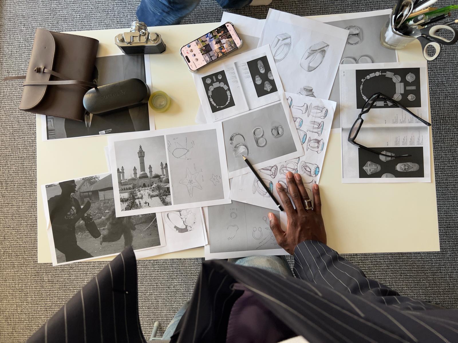Overhead view of a person sketching jewelry designs with reference images and tools on a table.