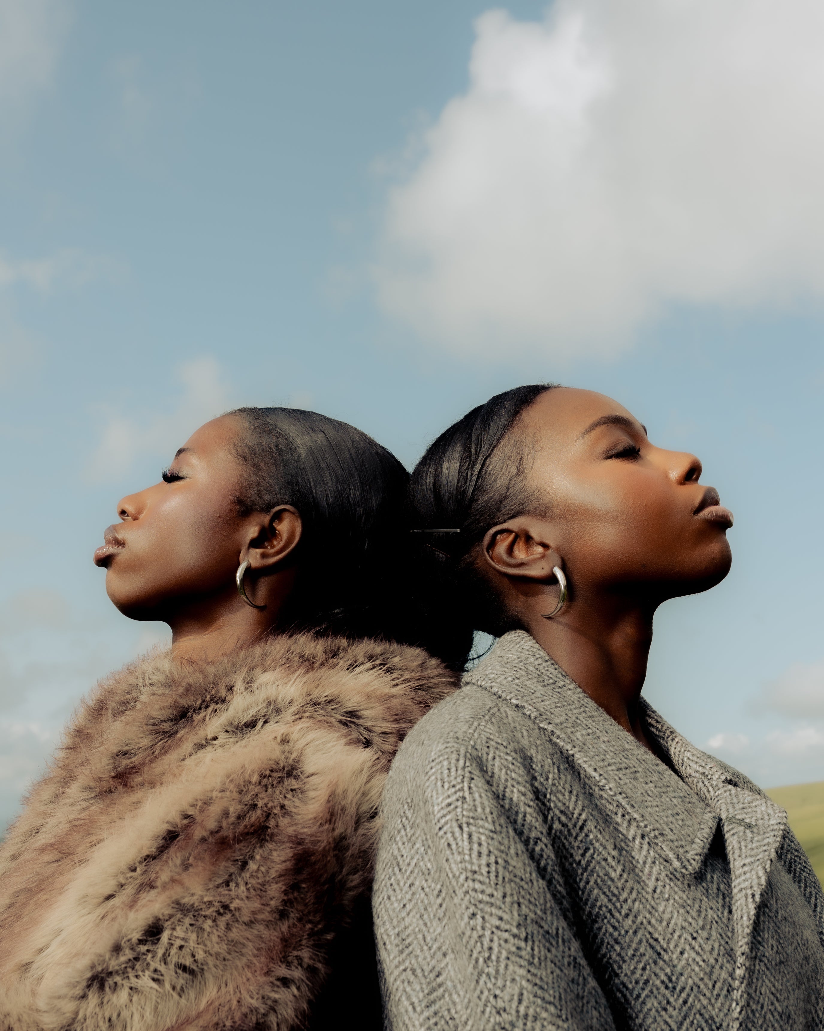 wo Senegalese sisters standing back-to-back in textured coats beneath a blue sky  Maison Nwar editorial shoot in Wales.