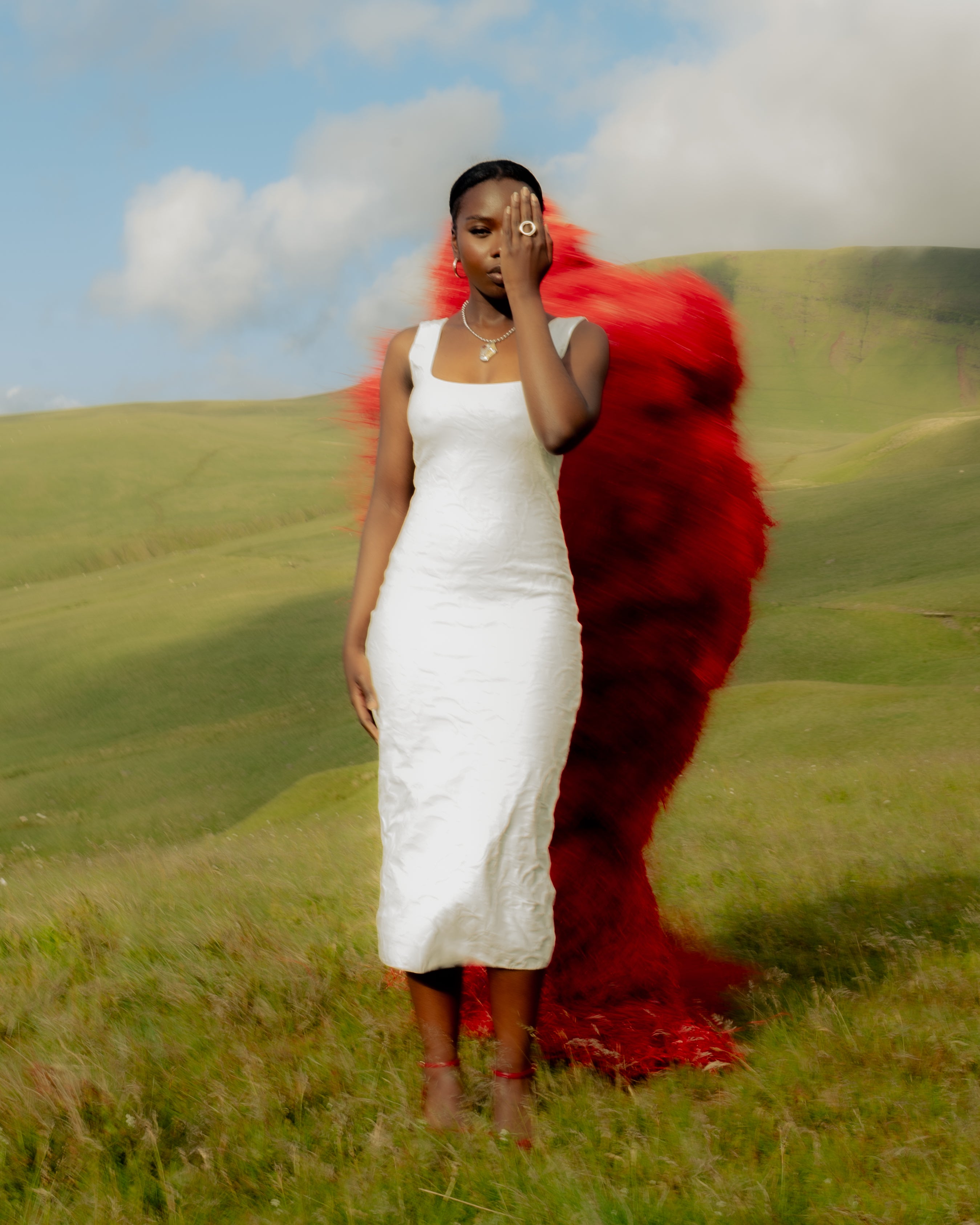 A Senegalese woman wearing Maison Nwar jewelry in a white dress, standing before the Kankurang figure in the Welsh hills  cultural editorial shoot.