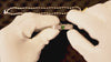 White-gloved hands holding and inspecting a silver ring with a small green stone over a dark surface, with a chain in the background.