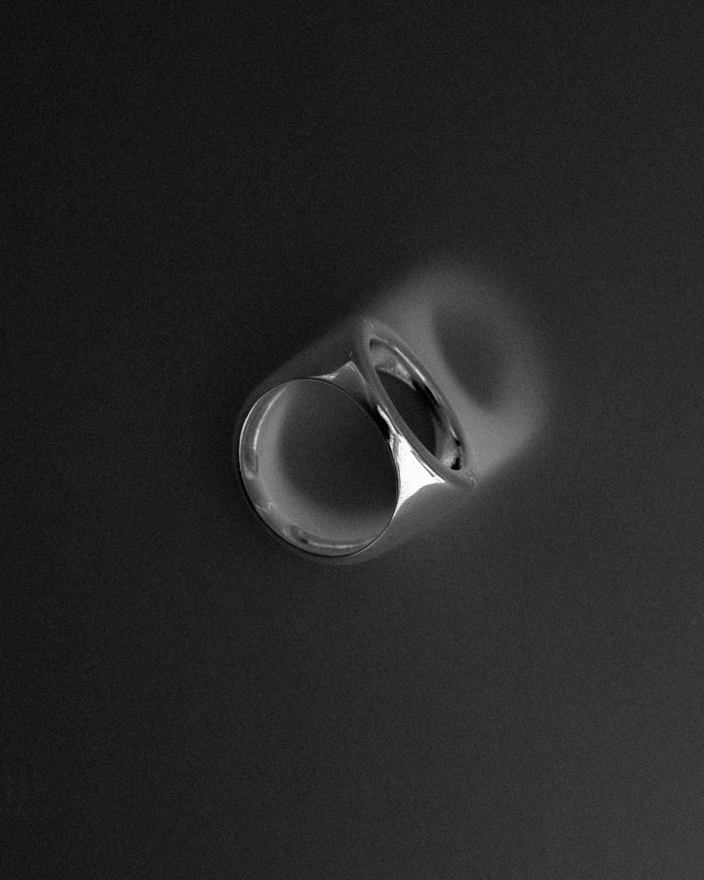 Minimalist silver signet ring photographed at an angle on a dark, softly blurred background with directional light creating a subtle glow and shadow gradient.